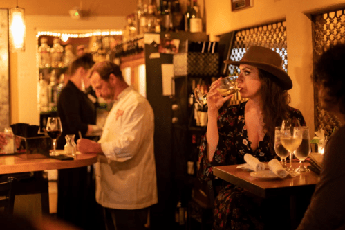 A woman in a hat sips a drink at a bar, while a chef and a server work in a cozy, warmly lit restaurant.